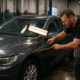 A technician performs paintless dent repair on a dark sedan with hail damage in a professional auto body shop, using specialized tools under bright lighting.